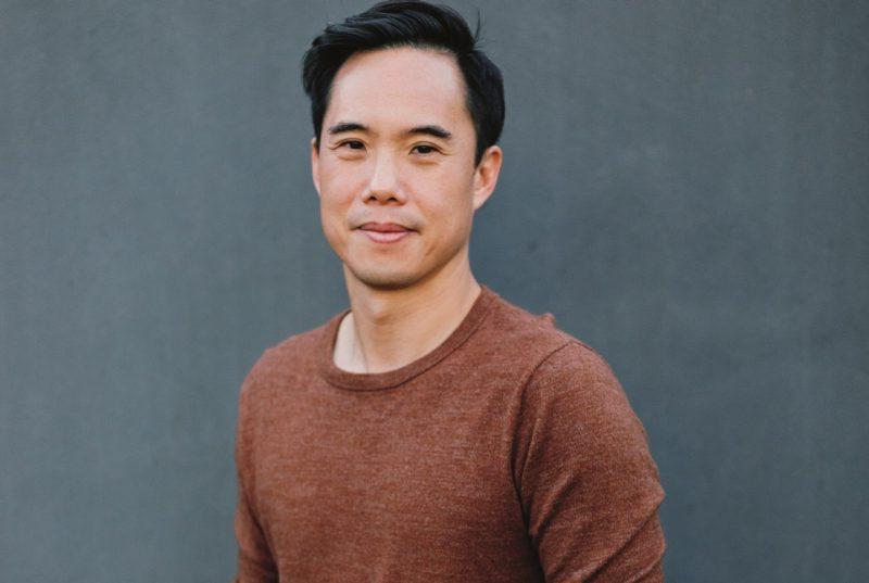 Author Charles Yu, a young Asian man with short dark hair, wears a rust brown T-shirt and smiles towards the camera in front of a medium grey background.