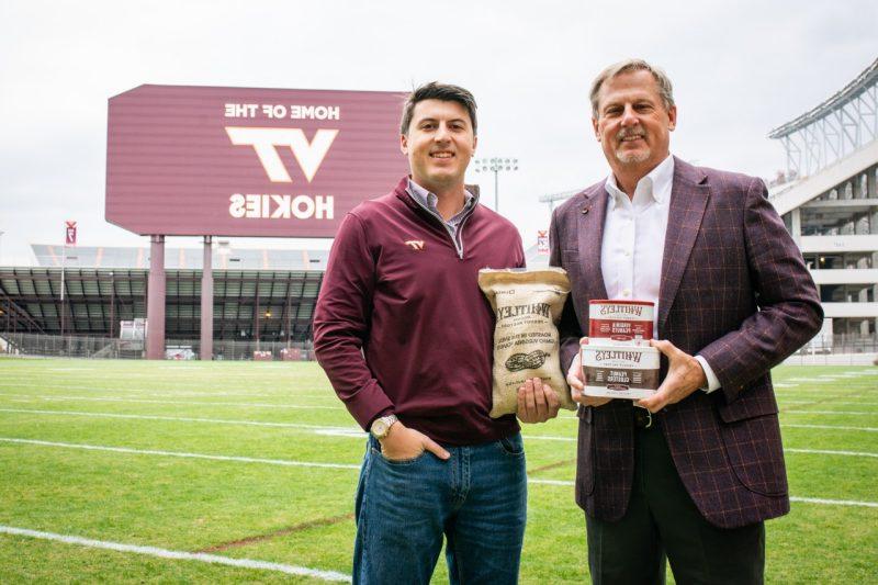 Two men stand on a football field in front of a sign holding food products.